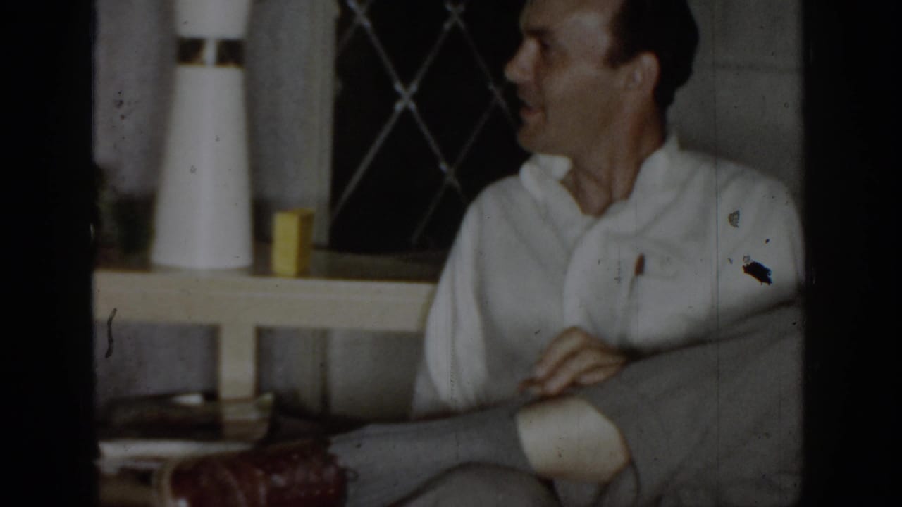 Man in white shirt seated on a grey sofa, smiling, with a lattice wall and cylindrical objects on a side table behind him.