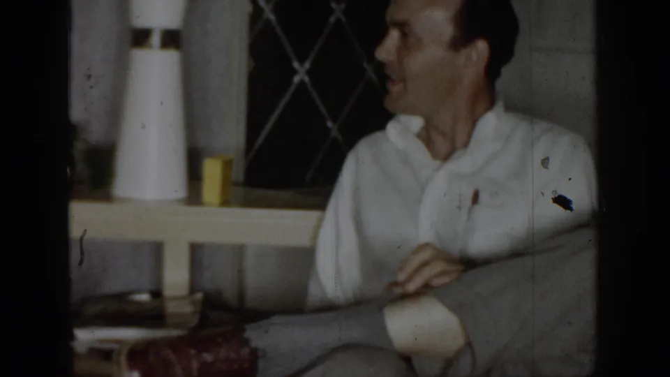 Man in white collared shirt relaxes on a grey sofa in a mid-1960s domestic interior; a lattice wall and white cylindrical objects on a side table are visible behind him.