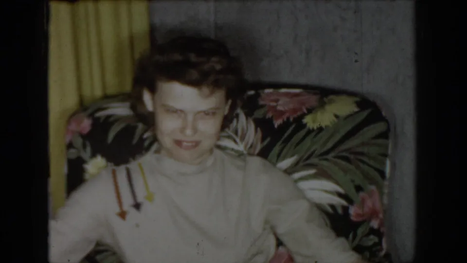 Woman in a light turtleneck with downward arrow graphic print seated in a dark floral armchair in a mid-1960s domestic interior, yellow curtains visible behind her.