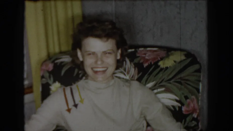 Woman in light turtleneck with arrow graphic print laughing openly in a dark floral armchair in a mid-1960s domestic interior, yellow curtains behind her.