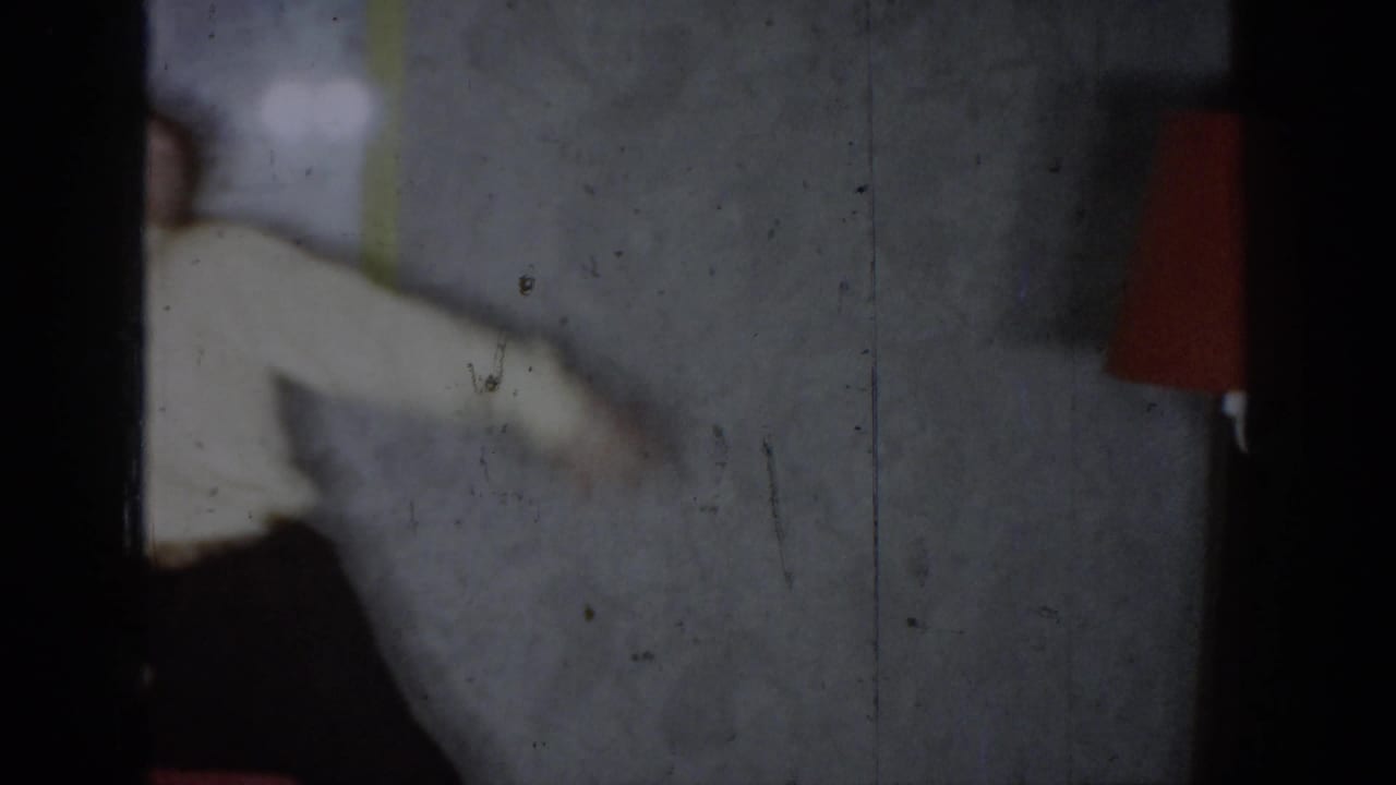 Partially visible figure in a white jacket with one arm extended, dark curtain at frame right, red object visible upper right.