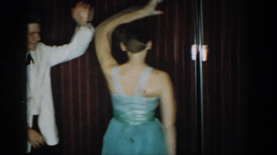 Teen couple mid-spin, man's arm raised and woman's bare back visible in light blue ballgown, against dark red curtain, 8mm home movie, 1965, Saint Louis, Missouri.