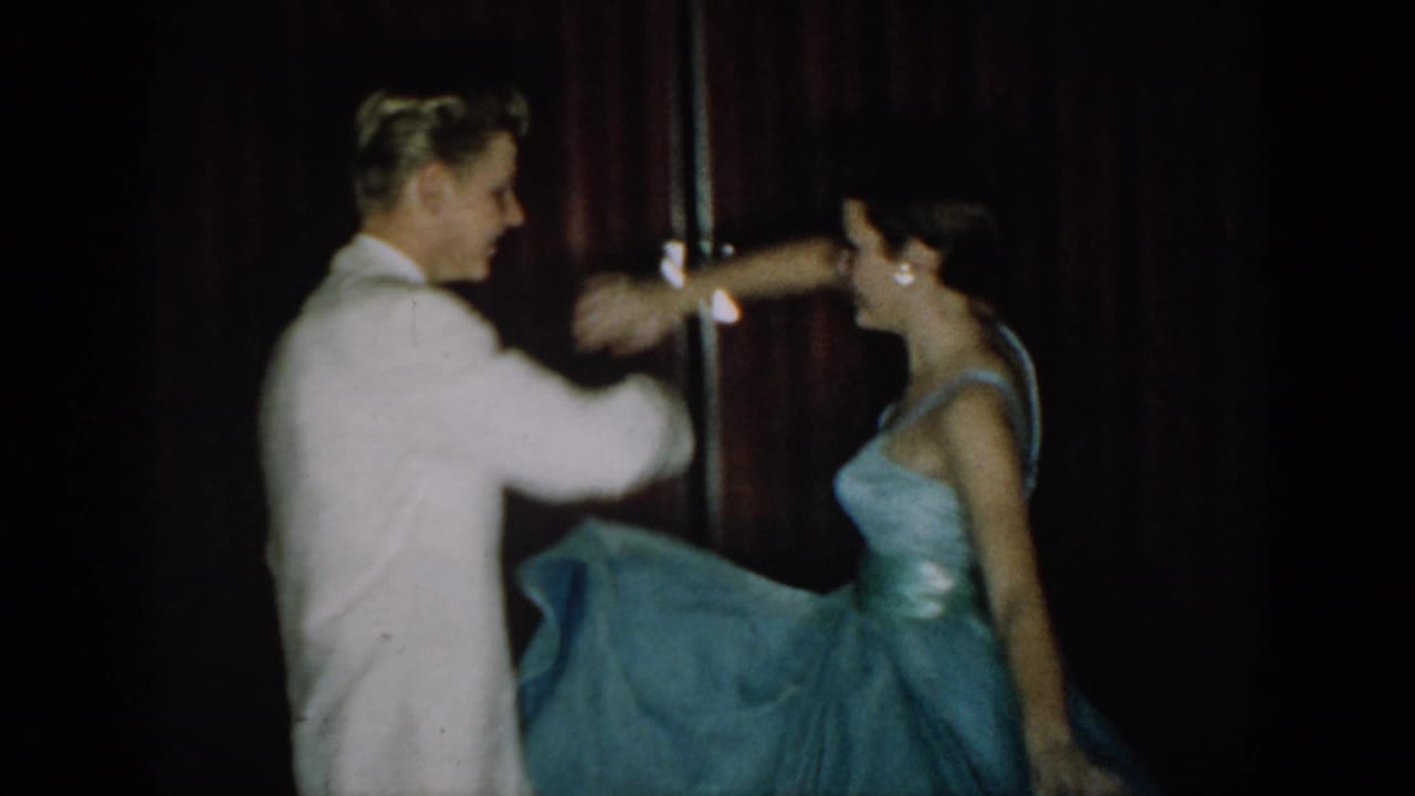 Young couple mid-dance turn at what appears to be a mid-1960s formal event, likely Saint Louis, Missouri; man in white dinner jacket, woman in light blue strapless ball gown, red velvet curtain behind.