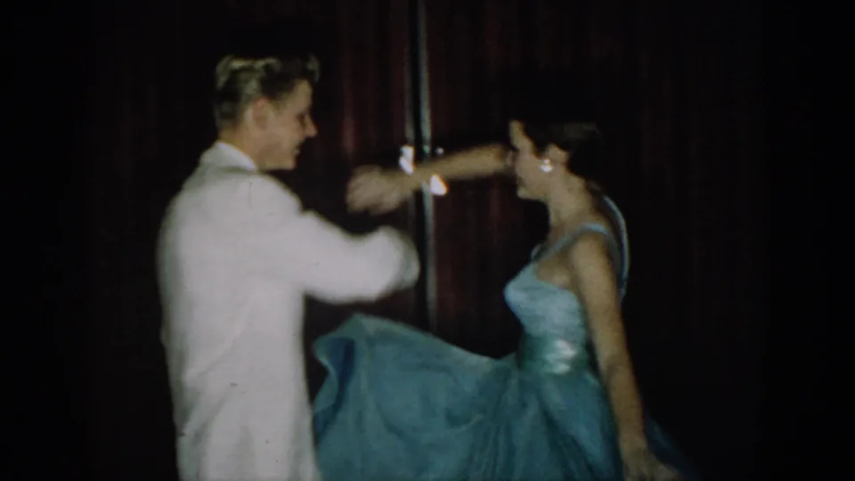 Young couple mid-dance turn at what appears to be a mid-1960s formal event, likely Saint Louis, Missouri; man in white dinner jacket, woman in light blue strapless ball gown, red velvet curtain behind.