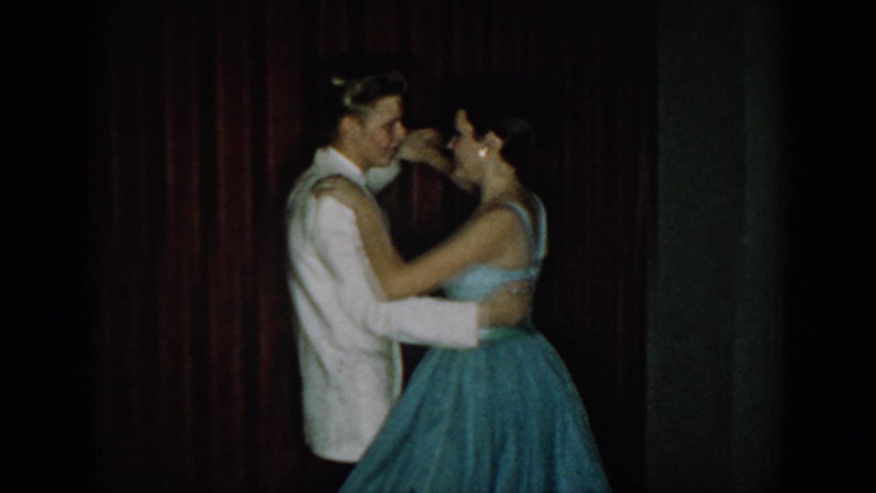 Young couple in close ballroom embrace at a mid-1960s formal event; man in white dinner jacket and woman in light blue full-skirted gown, dark red velvet curtain backdrop.