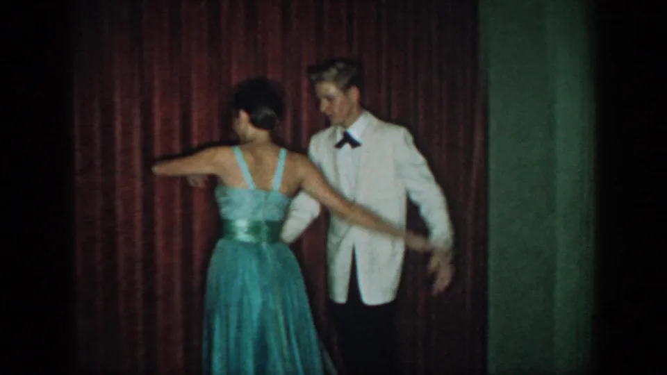 Rear view of couple at a 1960s formal dance event; woman's blue strapless gown shows low-back detail with wide sash, man in white dinner jacket and black bow tie, red velvet curtain behind.