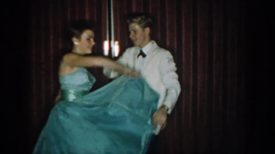 Couple at a mid-1960s formal event as woman's full blue ball gown skirt swirls from dance motion; man in white dinner jacket smiling, wall-mounted light sconce visible in background against red curtain.