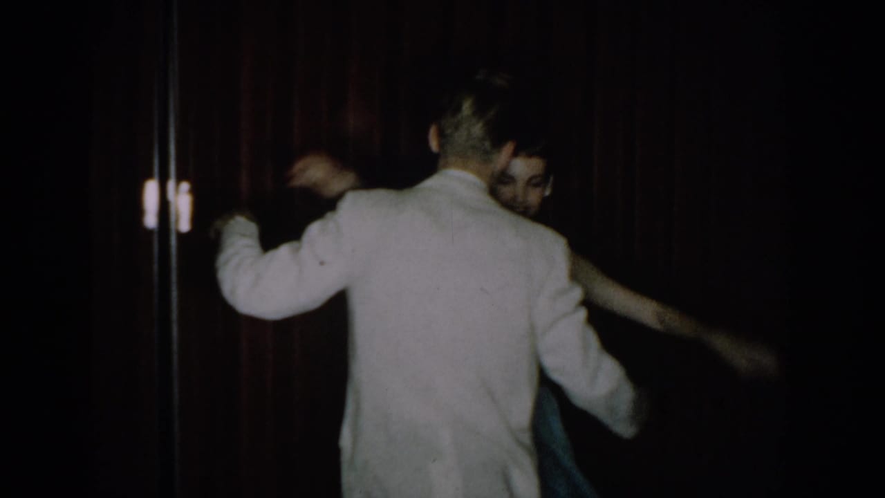 A man in a white dinner jacket and woman in a light blue gown dance viewed from behind at a mid-1960s formal party in Saint Louis, Missouri.