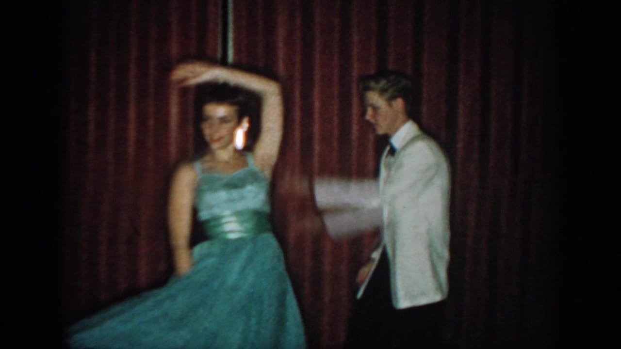 Woman in teal ball gown raising one arm in a spin turn; man in white dinner jacket watches, red curtain backdrop, 8mm home movie circa 1965.