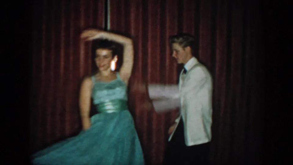 Woman in teal ball gown raising one arm in a spin turn; man in white dinner jacket watches, red curtain backdrop, 8mm home movie circa 1965.