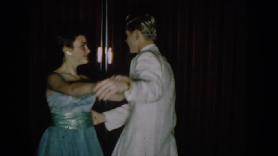 Couple in close hold, woman smiling toward camera; a hanging light fixture visible upper right, red curtain backdrop, 8mm home movie circa 1965 Saint Louis.