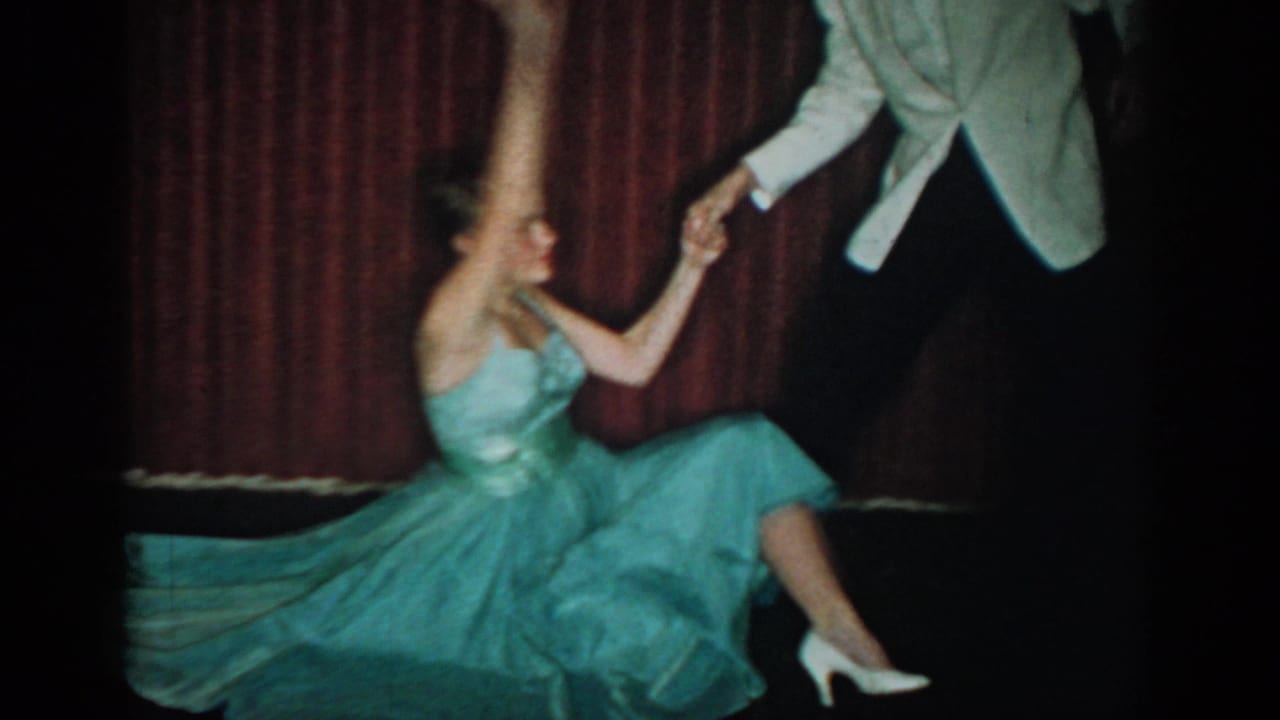 Young woman in teal ballgown mid-spin, held by man in white tuxedo jacket, against deep red curtain backdrop.