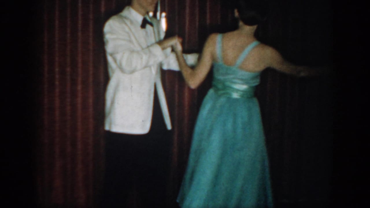 Couple in formal attire viewed from behind, woman in teal ballgown with back straps, man in white tuxedo with black bow tie, red curtain.