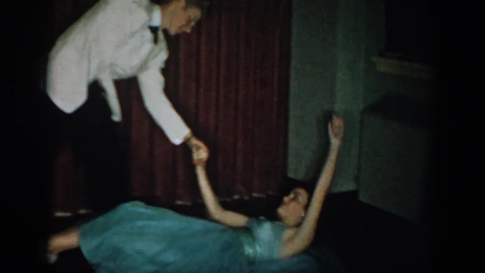 1965 home movie frame: woman in teal ballgown in dramatic floor-level dip, arm reaching up to partner in white tuxedo jacket bent forward, red curtain backdrop.