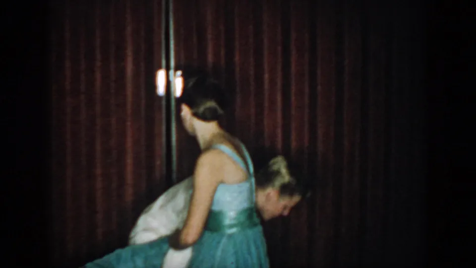 1965 8mm home movie frame of couple recovering from a dance dip: woman in teal ballgown, man in white tuxedo, small lamp or wall sconce visible behind them, red curtain.