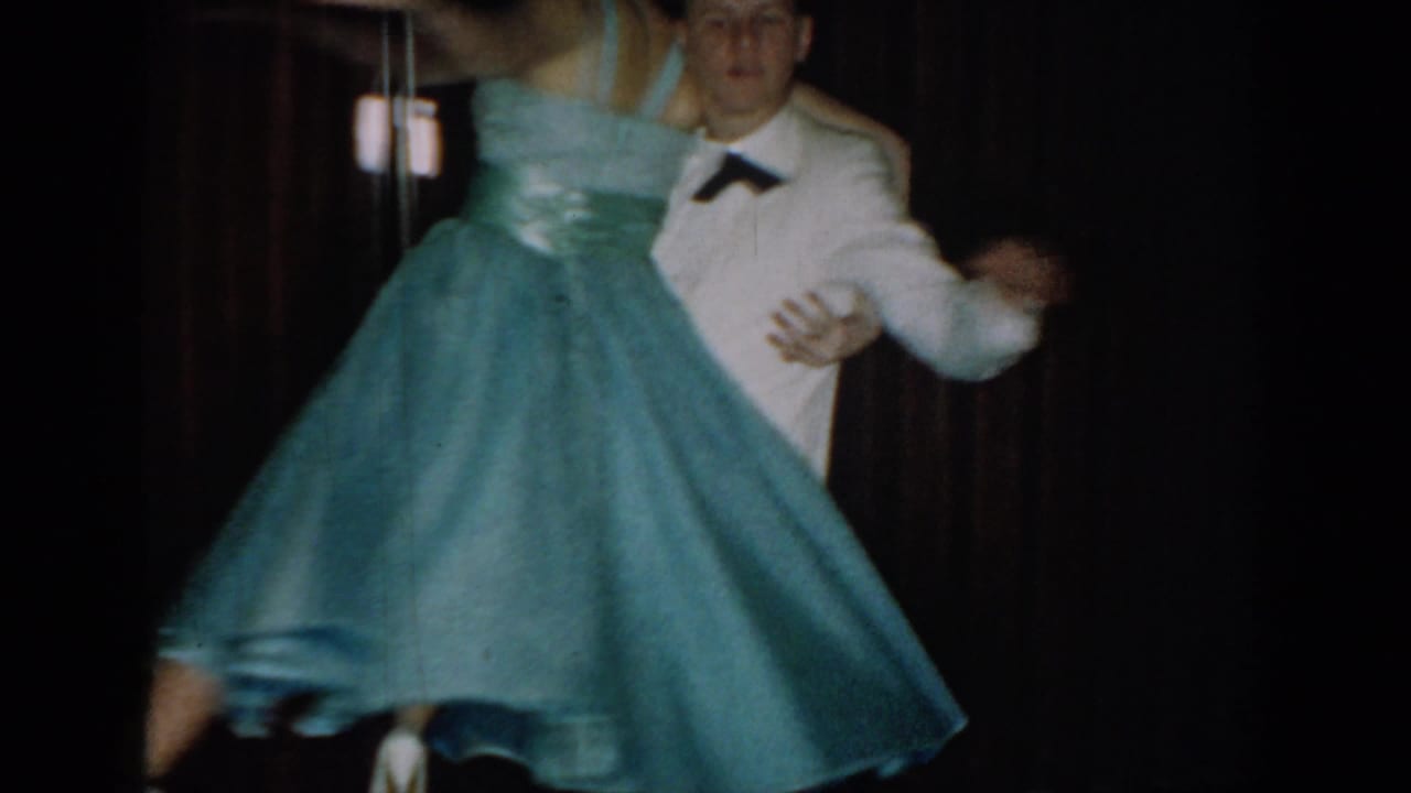 8mm home movie frame circa 1965 of a couple mid-dance, woman in full-circle blue formal gown billowing outward, man in white tuxedo jacket, dark backdrop.