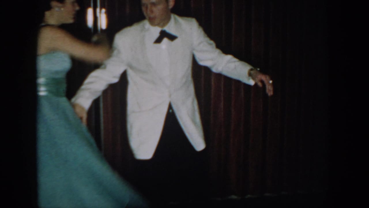 8mm frame circa 1965 showing a man in white dinner jacket and black bow tie with arms extended mid-dance, woman in full teal gown facing away, dark red curtain behind.