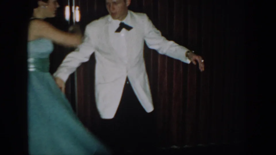 8mm frame circa 1965 showing a man in white dinner jacket and black bow tie with arms extended mid-dance, woman in full teal gown facing away, dark red curtain behind.