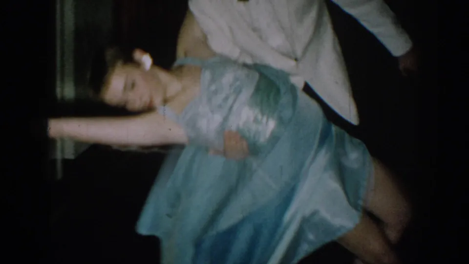 Tight 8mm frame circa 1965 capturing a low spin or dip mid-dance, woman's head near torso level, blue satin dress and white tuxedo jacket filling the frame with pronounced motion blur.