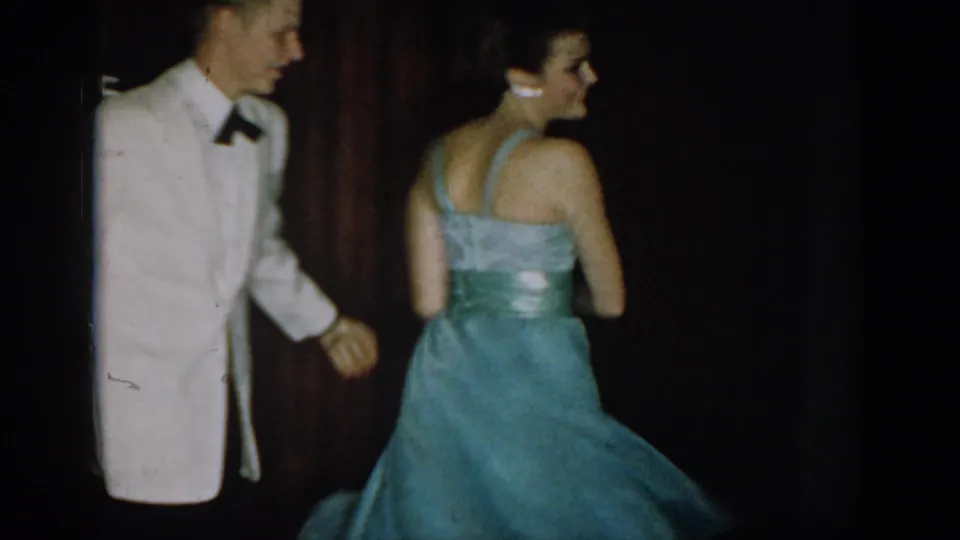 8mm frame circa 1965 showing a man in white dinner jacket smiling in profile and a woman from behind in a teal halter-neck formal gown with upswept dark hair, curtain backdrop.