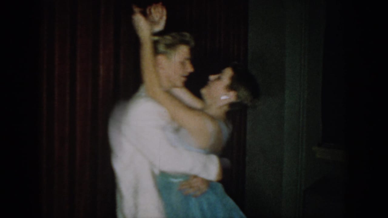 8mm home movie, circa 1965, Saint Louis, Missouri: young couple in formal attire mid-dance with arms raised against dark red velvet curtain.