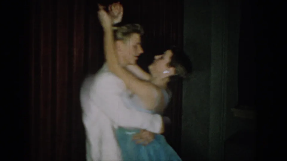 8mm home movie, circa 1965, Saint Louis, Missouri: young couple in formal attire mid-dance with arms raised against dark red velvet curtain.