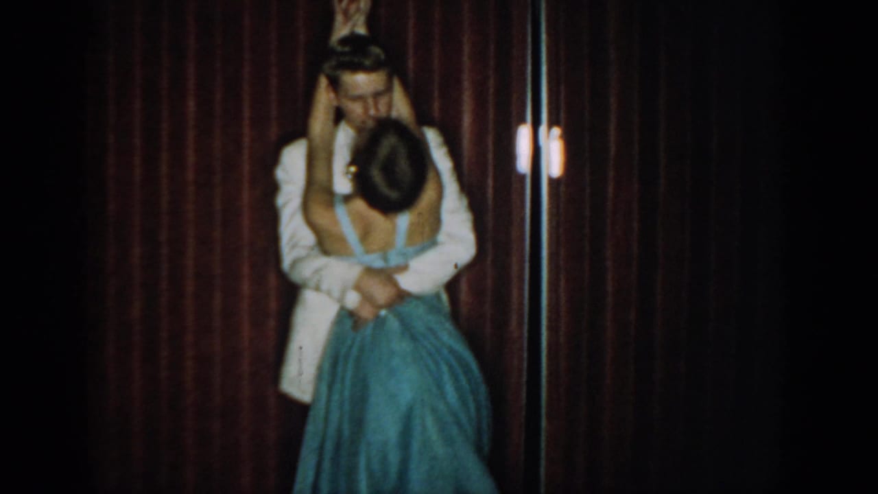 Man in white tuxedo embracing woman in blue gown from behind, heads close together, small light fixture visible at right edge.