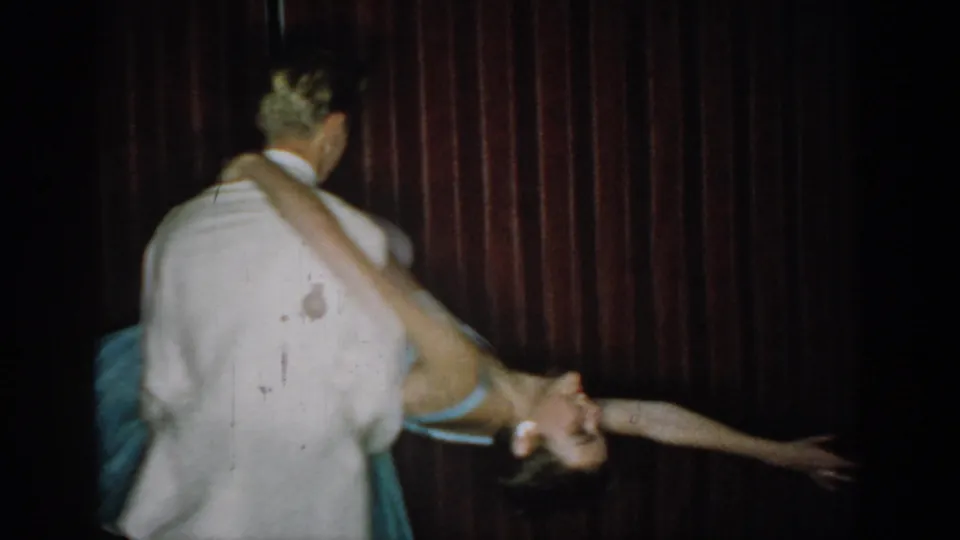 8mm home movie, circa 1965, Saint Louis: man in white tuxedo executes dramatic ballroom dip, woman's arm outstretched and blue gown trailing against dark red curtain.
