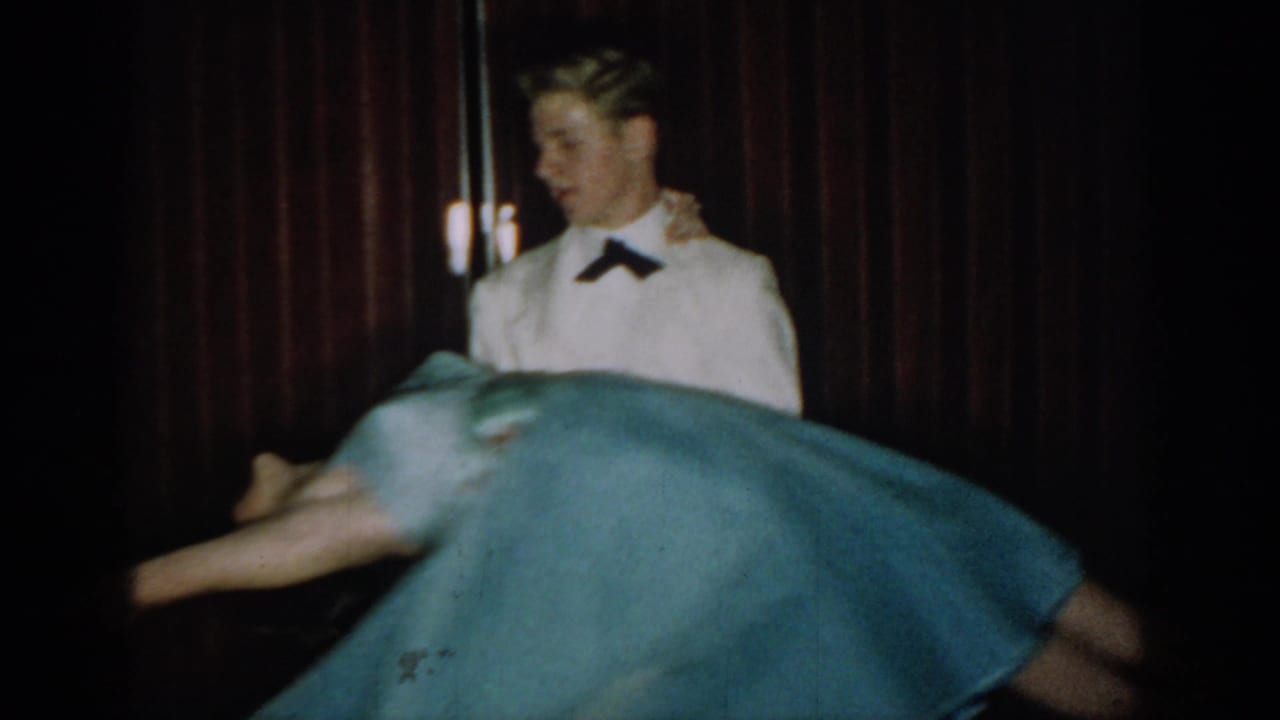 1965 8mm home movie frame: young man in white tuxedo with dark bow tie, woman's full-skirted light blue dress caught mid-spin, dark red curtain backdrop.