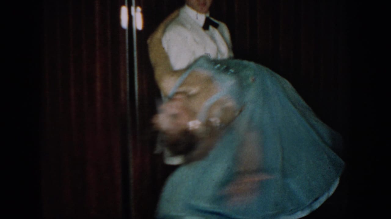 Couple close together mid-spin; man in white tuxedo jacket, woman's blue dress swirling dramatically against dark red curtain.