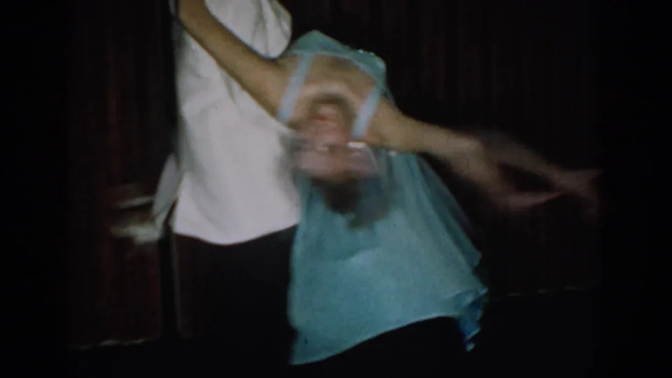 8mm home movie frame, c. 1965: young couple mid-dance lift, man in white tuxedo supporting woman in flowing blue dress, dark background.