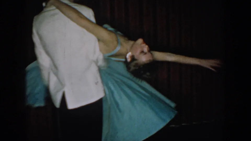 8mm home movie, c. 1965: woman in full-skirted blue dress dramatically dipped backward by man in white tuxedo jacket, dark red curtains in background.