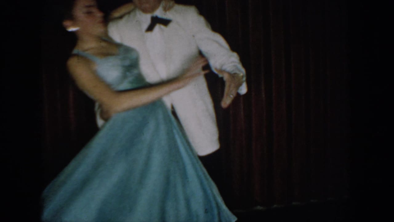 Man in white dinner jacket and woman in powder-blue ballgown dance in close hold before a deep red velvet curtain, circa mid-1960s.
