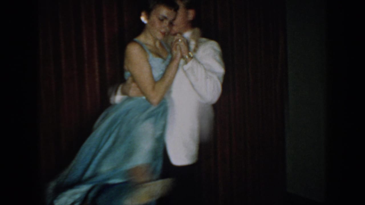 Dancing couple in tight embrace, the woman's full blue skirt prominent in the foreground; red curtain backdrop at a mid-1960s formal venue.