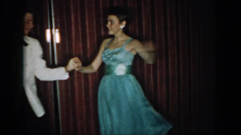 Full-figure view of dancing couple holding hands at arm's length, man in white jacket at left, woman in blue gown at right, stage sconce light and red curtain behind, circa mid-1960s.