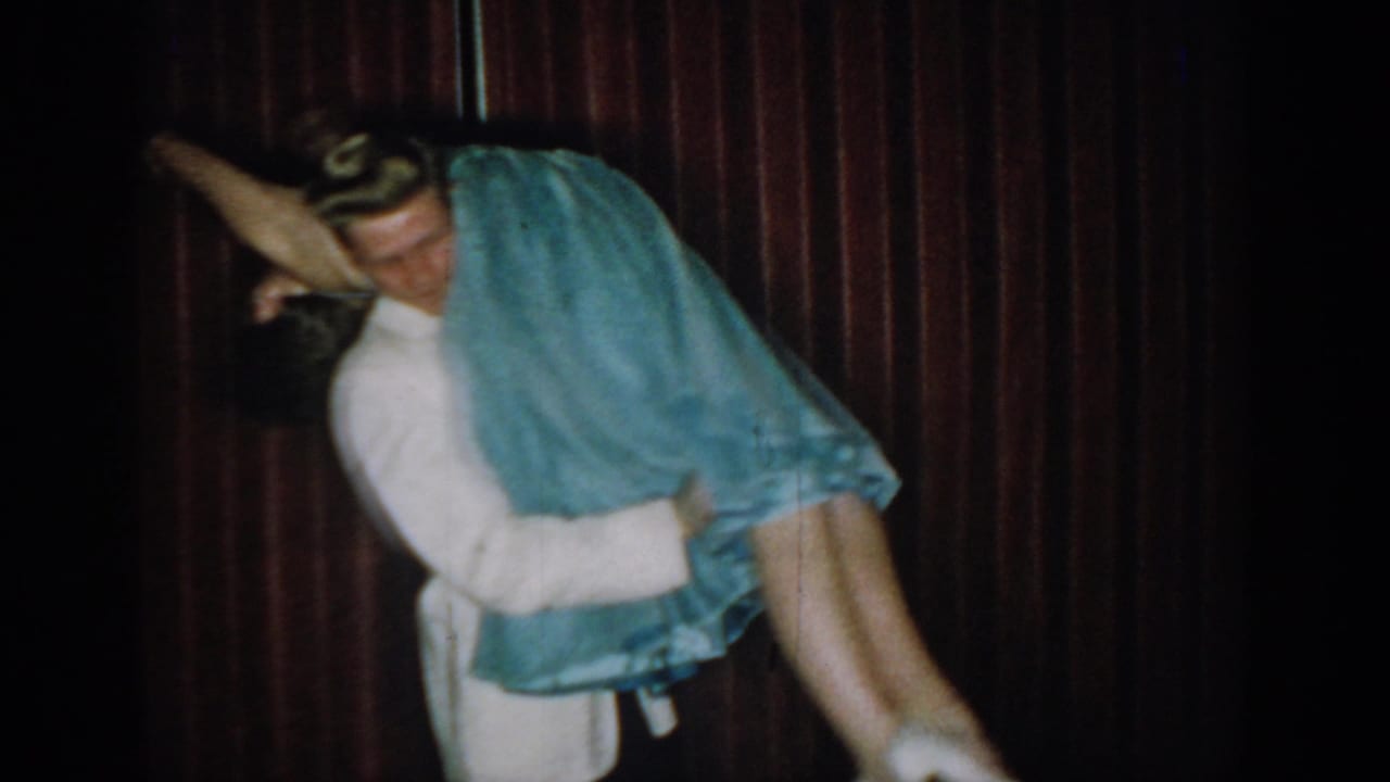 Mid-lift action captured on 8mm — man in white formal jacket raises woman in light blue ball gown, dress fabric swirling upward, against a dark red curtain, Saint Louis, circa 1965.