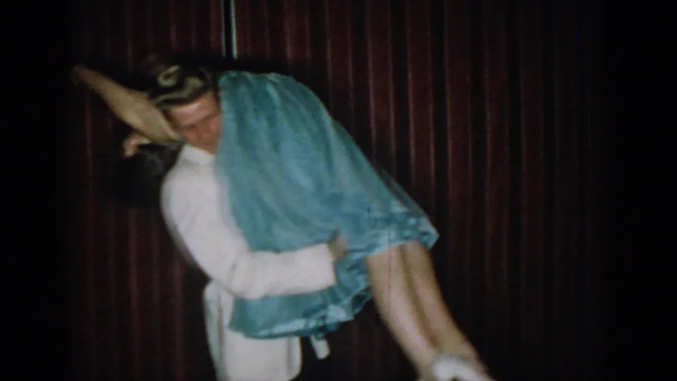 Mid-lift action captured on 8mm — man in white formal jacket raises woman in light blue ball gown, dress fabric swirling upward, against a dark red curtain, Saint Louis, circa 1965.