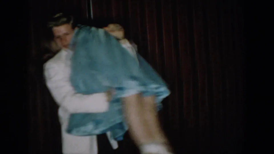 8mm home movie frame capturing a couple in energetic dance movement — man in white formal attire supports woman in blue ball gown, skirt fanning wide, against dark red curtain, circa 1965.