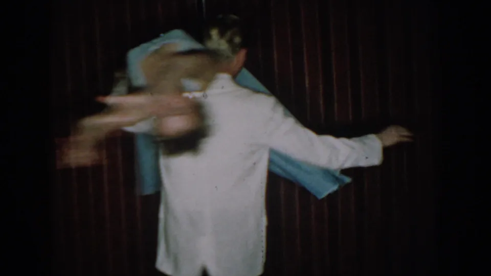 Final moments of 8mm dance sequence — woman in blue ball gown spins outward from man in white jacket, strong motion blur conveying speed, dark red curtain backdrop, Saint Louis, circa 1965.