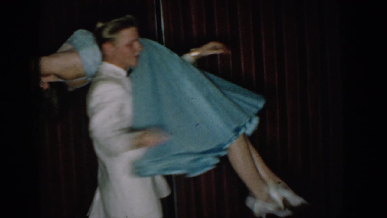 Man in white shirt and woman in light blue flared dress dancing against dark red curtain, dress billowing with motion.
