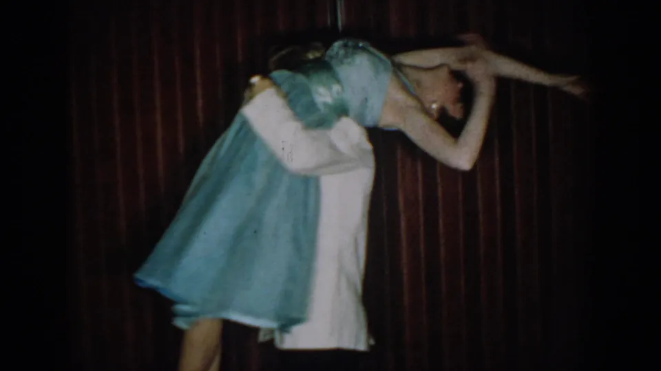 Man and woman in an expressive partnered dance pose against dark red curtain, woman bent forward with arms wide, circa 1965.