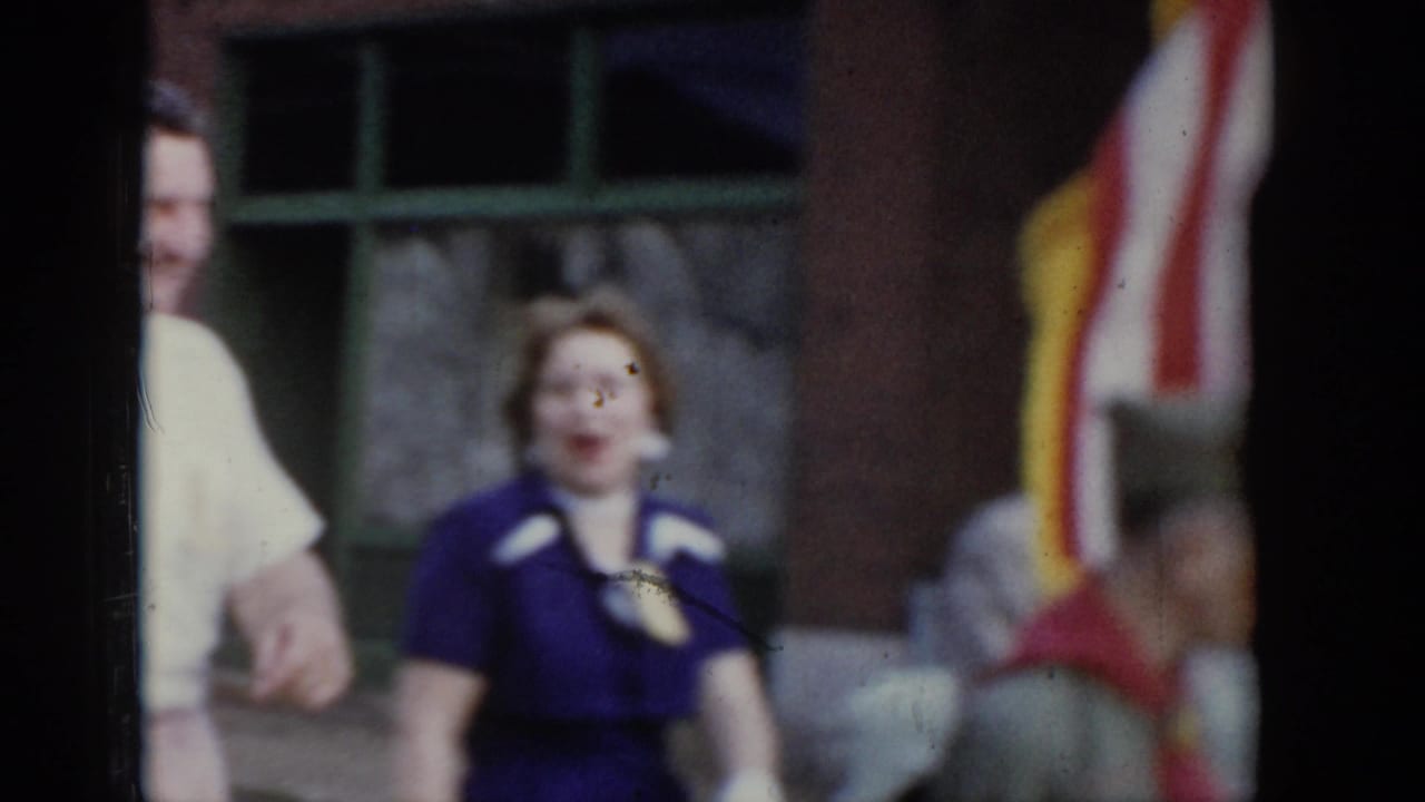 Smiling woman in purple dress with white collar and pin looking toward camera during a 1965 Saint Louis civic parade; American flag and brick building in background