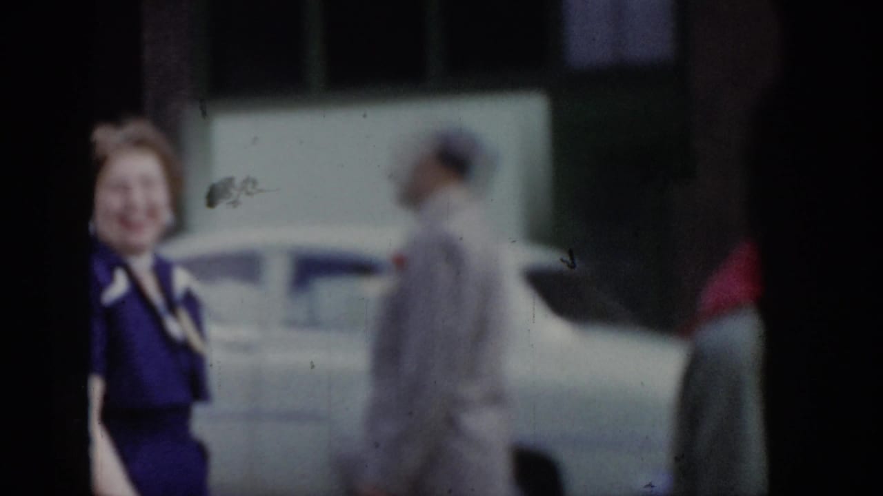 Woman in purple dress and man in light grey suit walking together on street, 1960s-era parked car visible in background