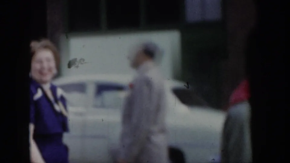 Woman in purple dress and man in light grey suit walking a 1965 Saint Louis parade route, with a period American automobile parked along the street
