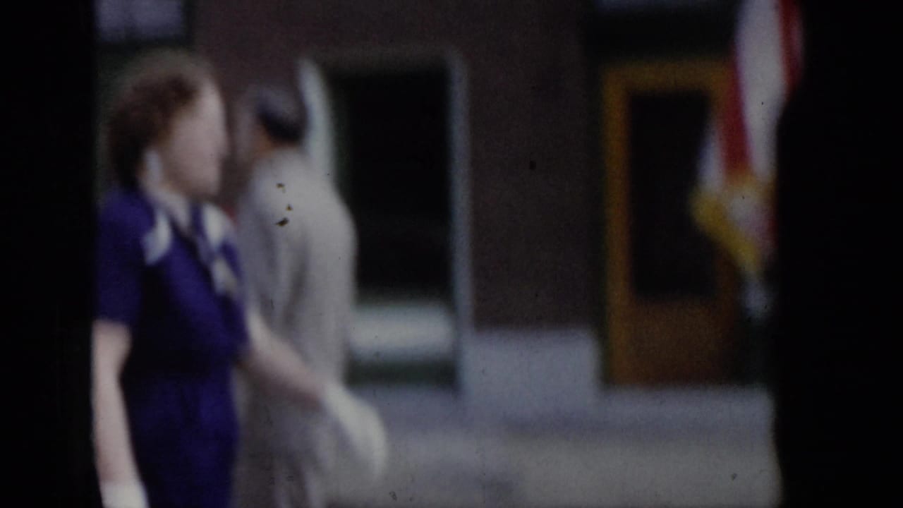 Close side view of woman in purple dress during a 1965 Saint Louis parade, with a companion in beige and a storefront with yellow door visible behind