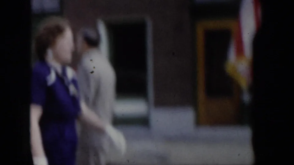 Close side view of woman in purple dress during a 1965 Saint Louis parade, with a companion in beige and a storefront with yellow door visible behind