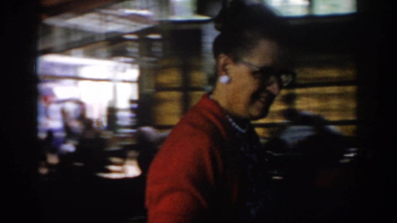 Woman in red cardigan and dark-framed glasses smiling in profile at a crowded indoor gathering.