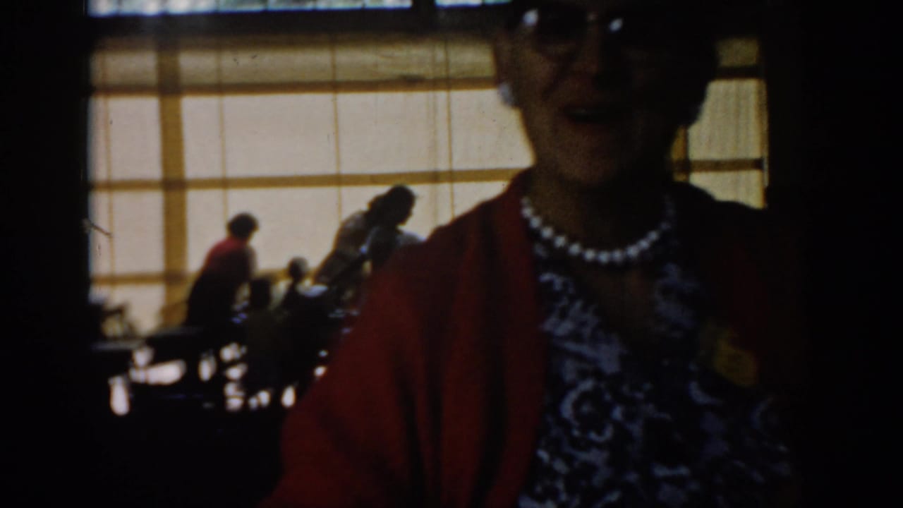 Torso close-up of woman in red cardigan and patterned dress with pearl necklace at indoor gathering; head partially cropped.