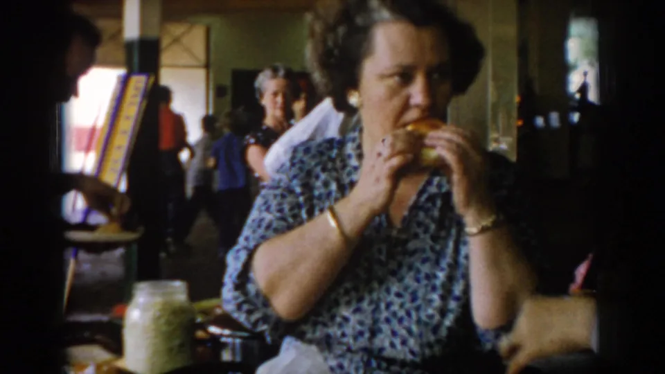A woman in a blue floral patterned dress and gold bracelet eats a roll or pastry at what appears to be a mid-1960s indoor community gathering; a glass jar and colorful multi-stripe banner are visible in the background.