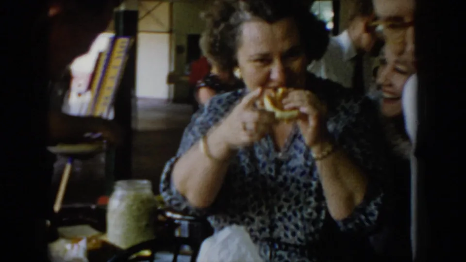 Continuation of shot showing a woman in a blue floral patterned dress eating at a 1960s indoor community event, with a colorful multi-stripe banner, a glass jar, and a man visible in the mid-ground.